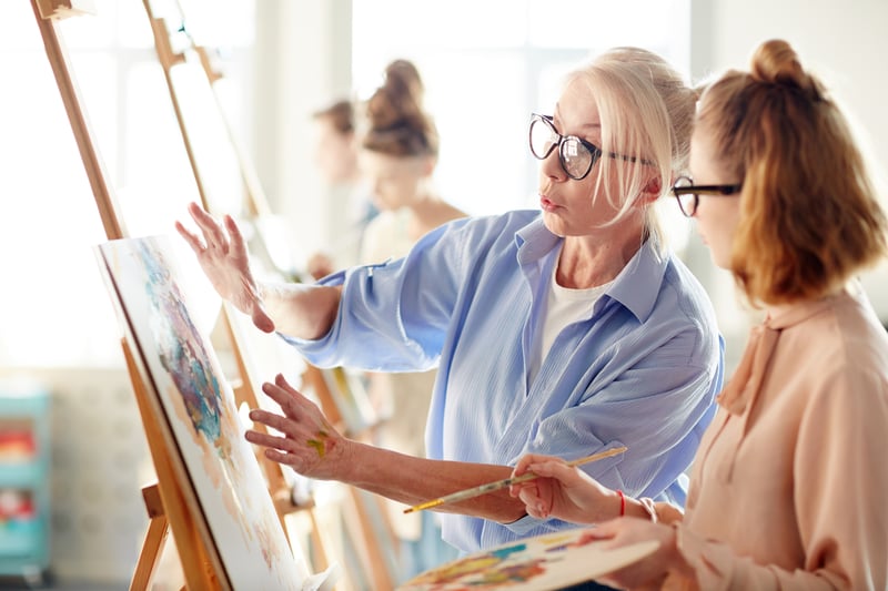 Woman wearing blue shirt and glasses - an art teacher directing an art student working at an easel in the art studio on an around the world cruise ship